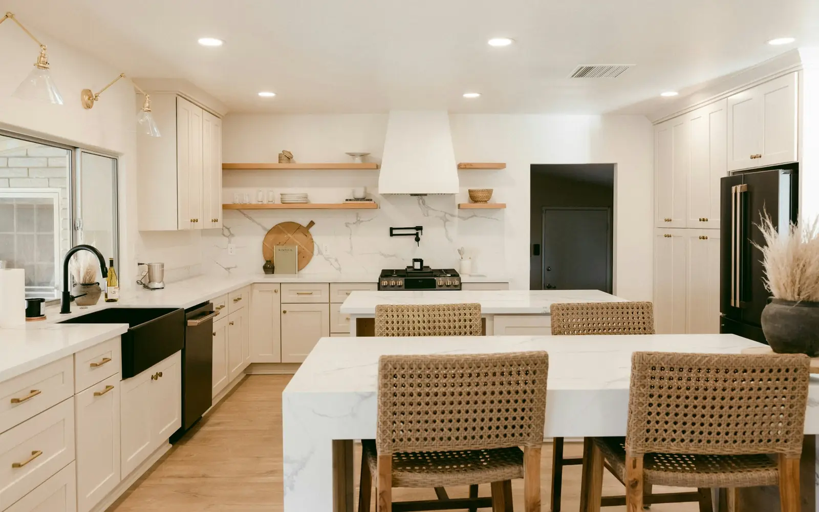 White shaker kitchen with two quartz islands, woven stools, a black farmhouse sink and open wood shelves