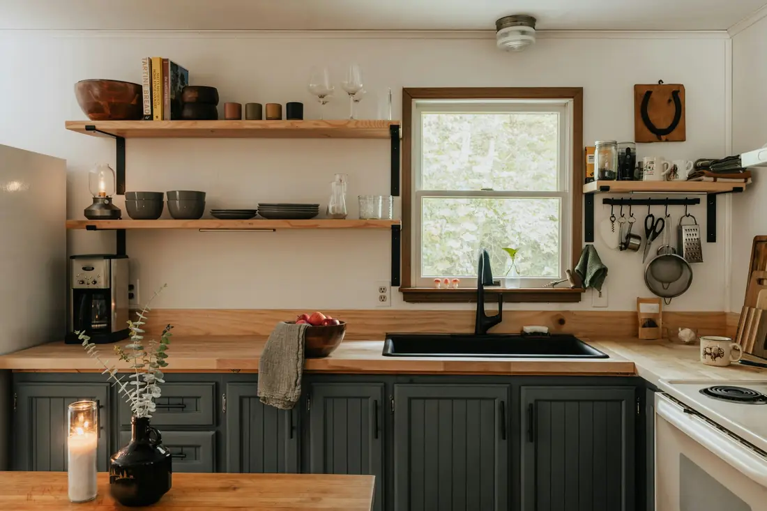 Rustic kitchen with butcher block counters, gray painted cabinets, open wood shelves and a black sink under a window