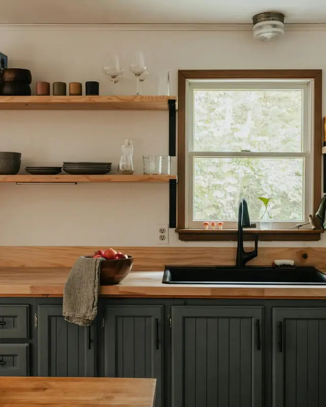 Rustic kitchen with butcher block counters, gray painted cabinets, open wood shelves and a black sink under a window