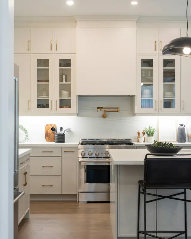 White shaker kitchen with glass-front upper cabinets, brass pulls and a pot filler over a gas range