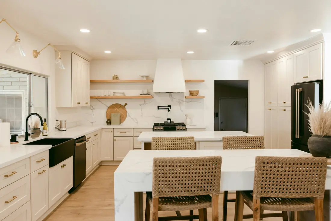White shaker kitchen with two quartz islands, woven stools, a black farmhouse sink and open wood shelves