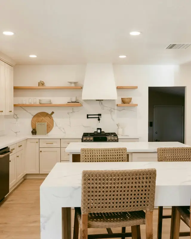White shaker kitchen with two quartz islands, woven stools, a black farmhouse sink and open wood shelves